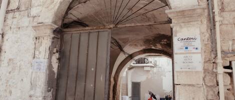 An arched entrance to a building with a closed metal gate, a sign for 'Cantovi ARCO ALTO ROOMS,' and a red scooter parked inside.