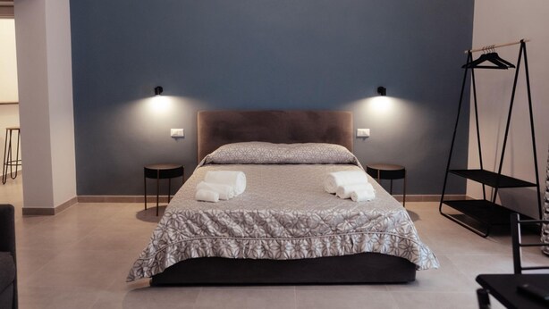 A neatly made bed with a patterned bedspread and two rolled towels, two bedside lamps, a black hanger stand, and a small table with a chair. - GuestHost - Guest Host - Elegant Studio Apt Maison Vittoria (Bari)