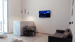 A modern hotel room with a bed, a sofa, a dining table with chairs, a TV mounted on the wall, and a large window. - GuestHost - Guest Host - Elegant Studio Apt Maison Vittoria (Bari)