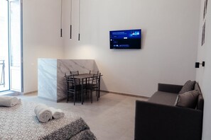 Ein modernes Hotelzimmer mit Bett, einer Couch, einem Esstisch mit Stühlen, einem an der Wand befestigten Fernseher und einem großen Fenster.
