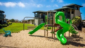 Ein Spielplatz mit Rutsche und Schaukeln, einem Sandbereich und einem Gebäude mit Balkon im Hintergrund.