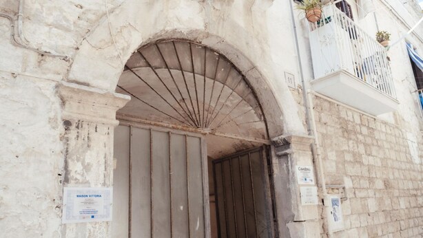 Entrada de edifício com uma grande porta em arco, um portão de metal e uma varanda com plantas em vasos.