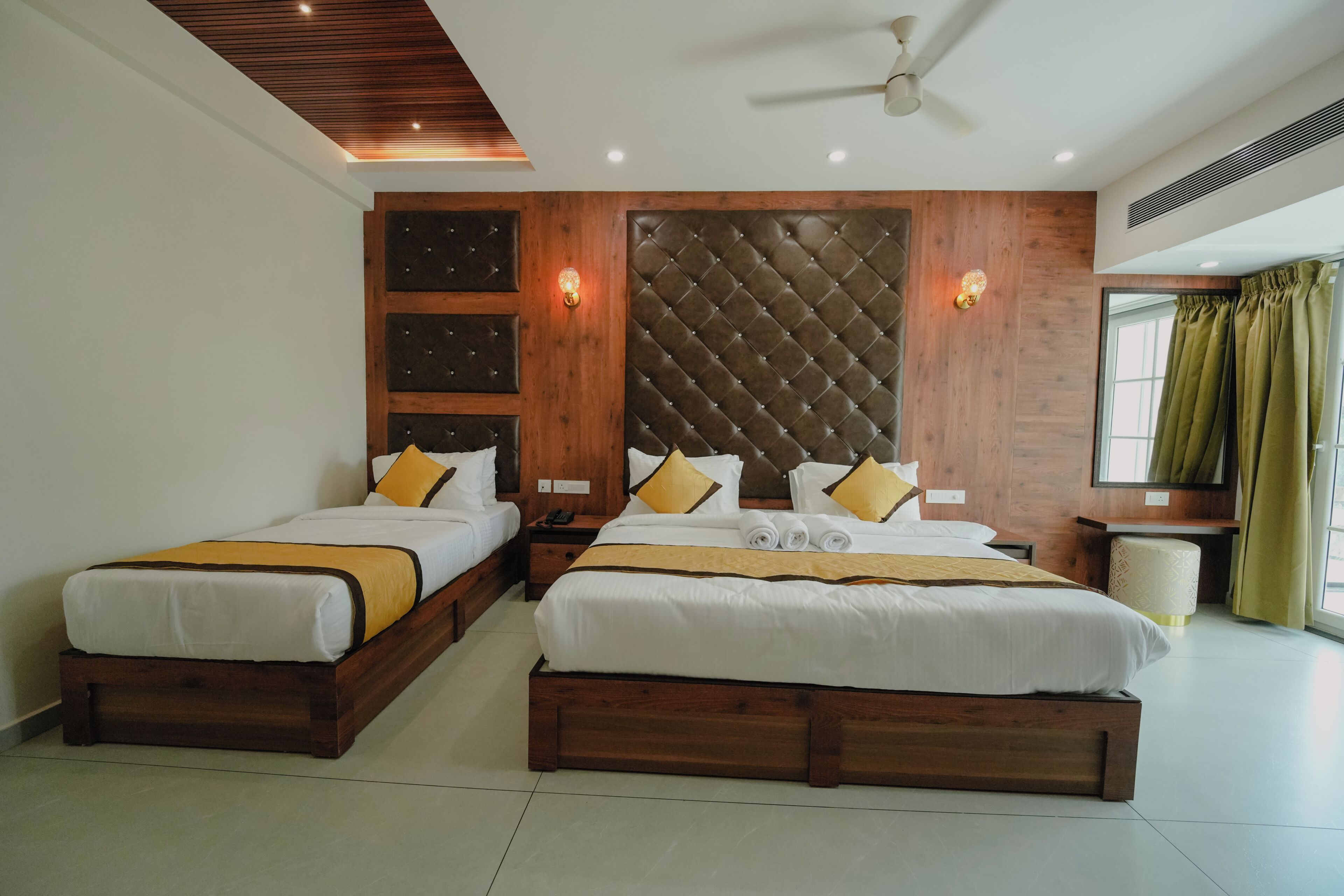 Habitación de hotel con dos camas, cabecera de madera, ventilador de techo y ventana con cortinas.