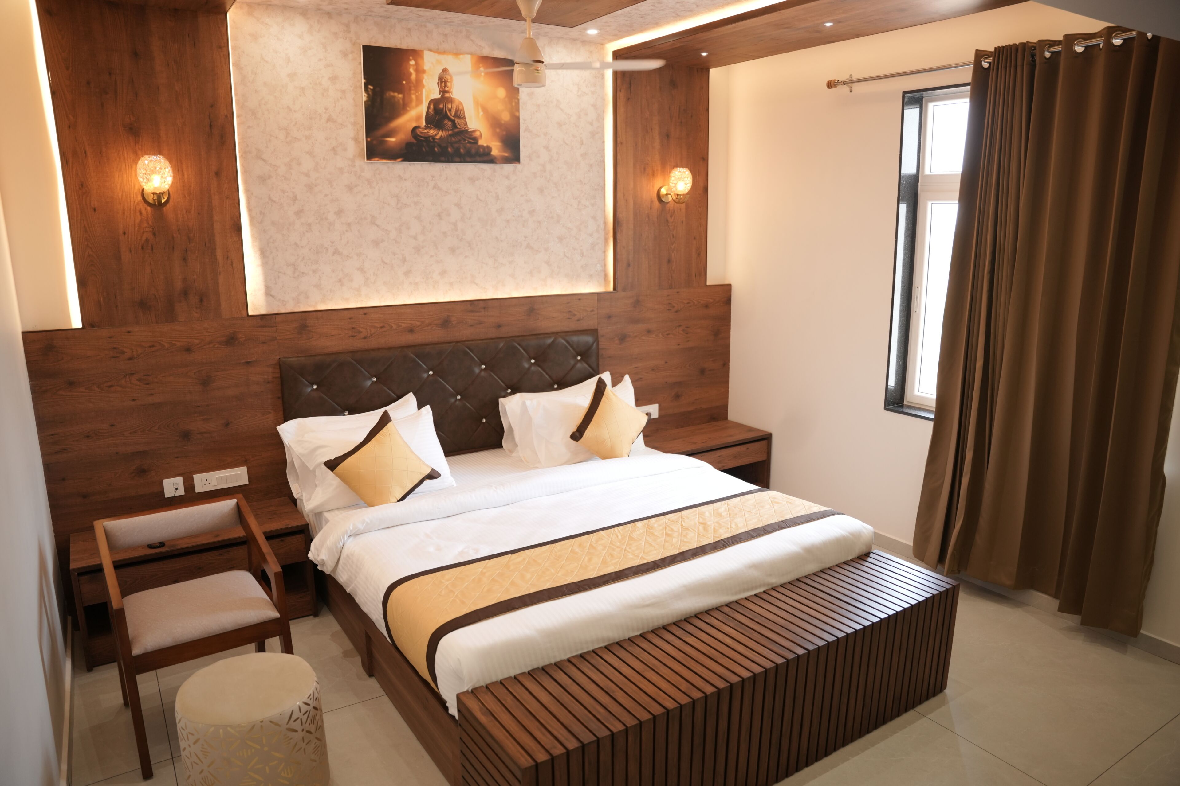 A modern bedroom with a wooden headboard, a large bed with white bedding, a bedside table, a bench, and a wall-mounted light fixture.
