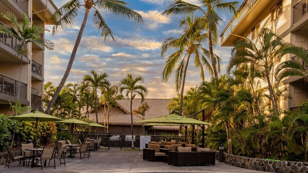 A swimming pool with lounge chairs and umbrellas, surrounded by palm trees and a building with balconies. - Two 2BR Condos, Pool, Hot Tub, Prime location (Kailua-Kona)