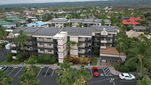 A multi-story building with solar panels on the roof, surrounded by greenery and a parking lot with cars. - Two 2BR Condos, Pool, Hot Tub, Prime location (Kailua-Kona)