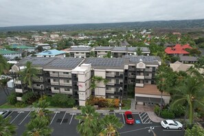 A multi-story building with solar panels on the roof, surrounded by greenery and a parking lot with cars. - Two 2BR Condos, Pool, Hot Tub, Prime location (Kailua-Kona)