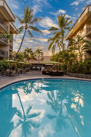 A swimming pool with lounge chairs and umbrellas, surrounded by palm trees and a building with balconies. - 2BR on Ironman Run Route, Pool & Hot Tub Access (Kailua-Kona)