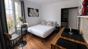 A compact hotel room with a bed, a small dining table, a kitchenette, and a view through large windows. - Studio Charmant Paris 15eme Proche Metro (Paris)
