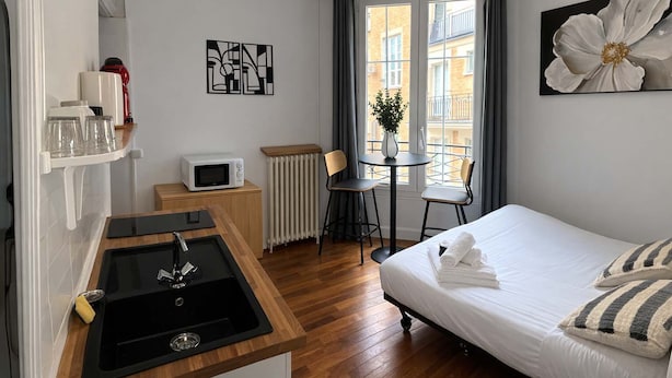 A hotel room with a kitchenette, a bed with pillows, a dining table with chairs, a radiator, and artwork on the walls. - Studio Charmant Paris 15eme Proche Metro (Paris)