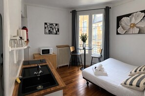 Une chambre d’hôtel équipée d’une kitchenette, d’un lit avec des oreillers, d’une table à manger avec des chaises, d’un radiateur et de tableaux décoratifs aux murs.