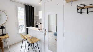 Uma cozinha compacta com bancada branca, uma geladeira pequena e um micro-ondas. Há dois bancos de bar de vime e uma porta branca com um espelho.