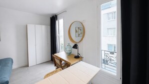 Quarto com mesa de madeira, uma cadeira, escrivaninha, guarda-roupa, janela com vista para edifícios e varanda.