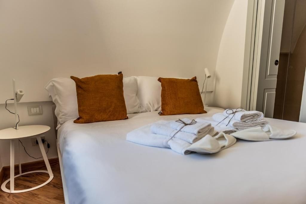 A neatly made bed with white and brown pillows, a small white side table with a power cord, and a set of towels and slippers on the bed.