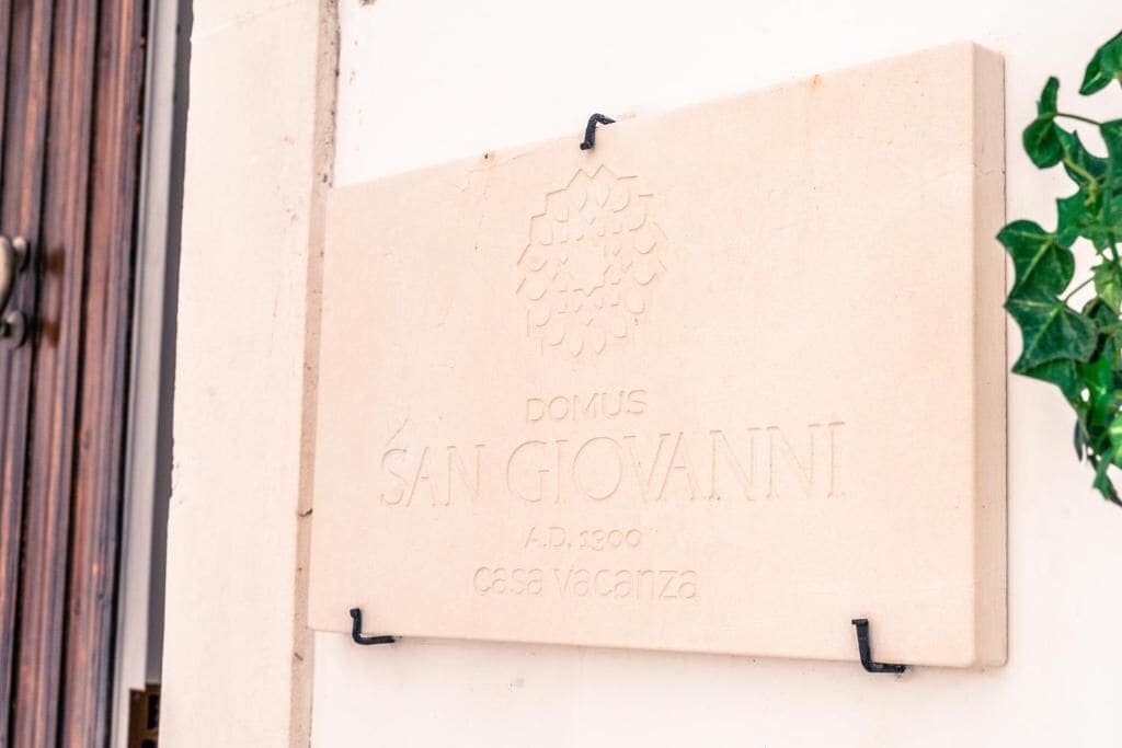 Eine Tafel mit der Inschrift 'DOMUS SAN GIOVANNI A.D. 1300 casa vacanza' an einer Wand befestigt.