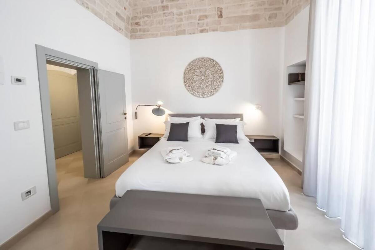 A hotel room with a bed, bedside tables, a wall-mounted lamp, a circular decorative item, and a door leading to another room.