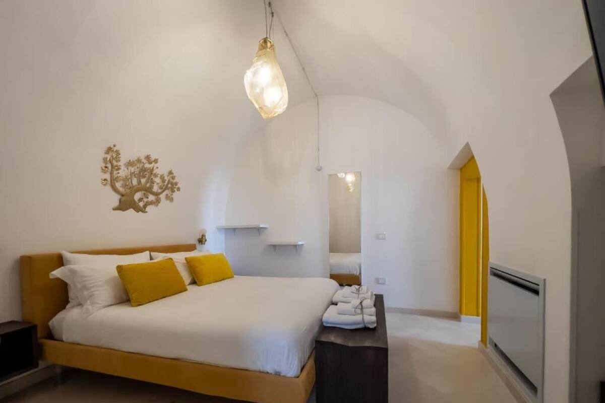 A bedroom with a bed, a bedside table, a hanging light fixture, a wall decoration, and a door leading to another room.