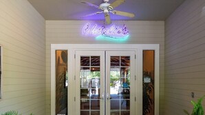 Ein Gebäudeeingang mit einer Neonreklame mit dem Schriftzug 'Charlie's' über einer Doppeltür, einem Deckenventilator und Topfpflanzen.
