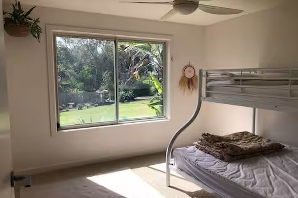 A room with a bunk bed, a large window with a view of greenery, a ceiling fan, and a dreamcatcher on the wall.