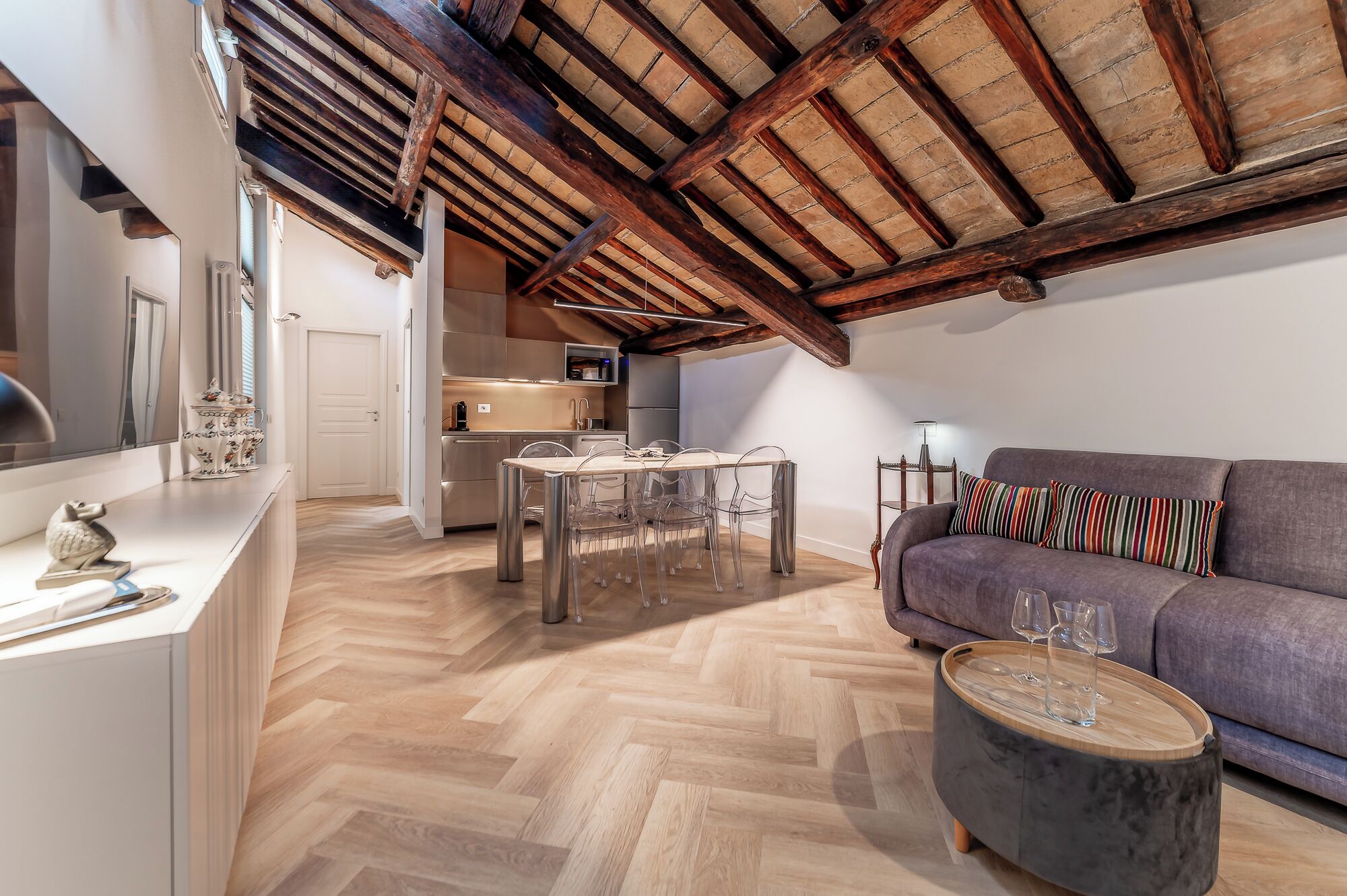 A modern loft apartment with exposed wooden beams, a herringbone floor, a kitchen with stainless steel appliances, a dining area with a glass table, and a gray sofa with striped pillows.