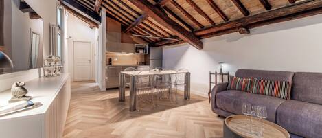 A modern loft apartment with exposed wooden beams, a herringbone floor, a kitchen with stainless steel appliances, a dining area with a glass table, and a gray sofa with striped pillows.