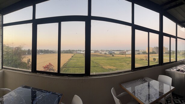 Ein Zimmer mit einem großen Fenster, das Blick auf Felder und Gebäude gewährt. Es steht ein Tisch mit Marmorplatte und ein Stuhl darin.