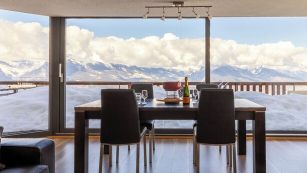 A dining area with a mountain view, a table set with chairs, and a bottle of champagne. - Hauts de Nendaz E by Interhome (Nendaz)