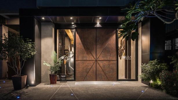 A modern hotel entrance with a large metal door, potted plants, and a well-lit pathway.