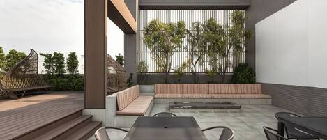 Un patio al aire libre con una mesa y sillas, un área de estar con cojines y vistas a árboles y edificios.