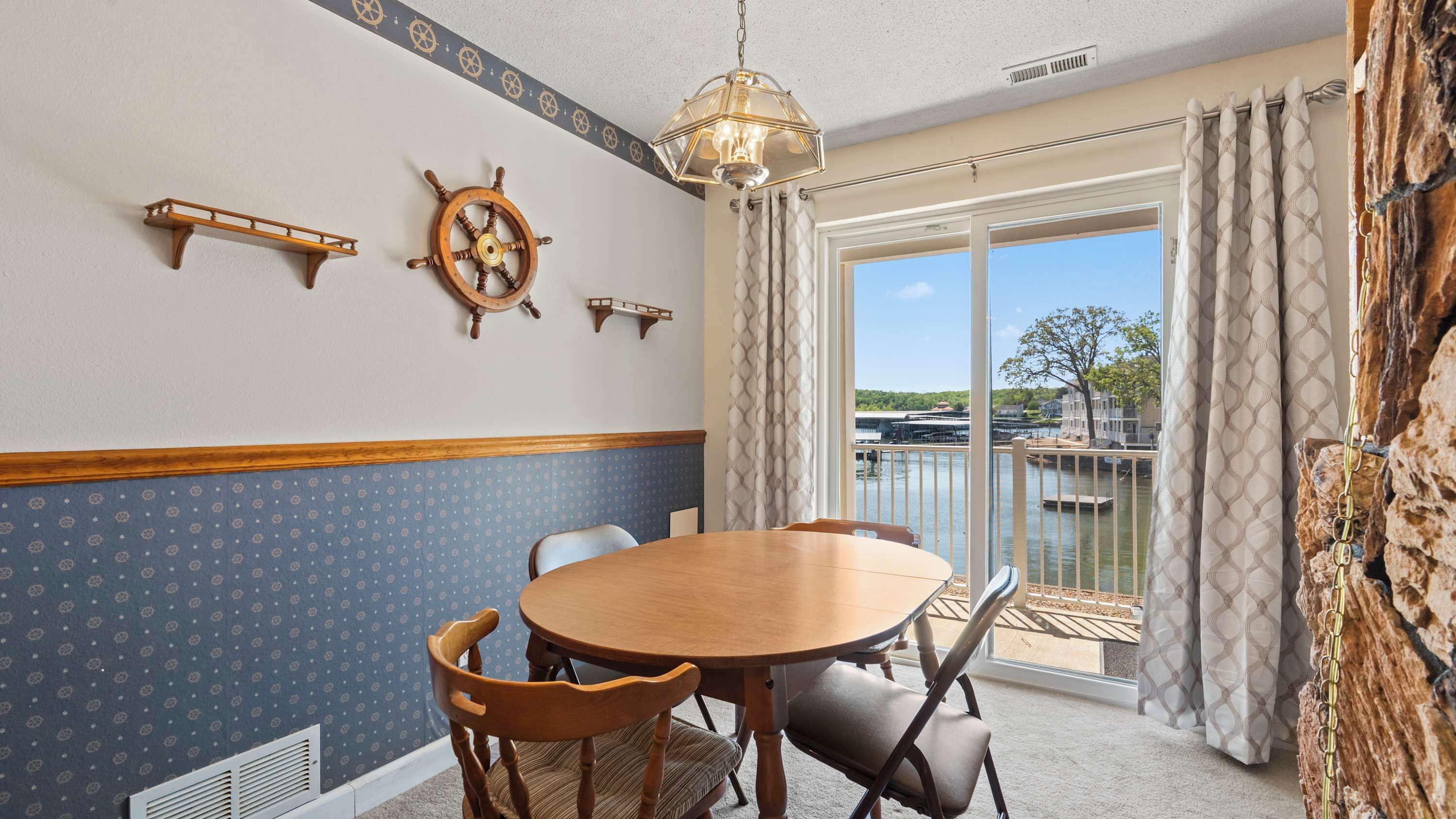 Un coin repas avec une table ronde, des chaises, un élément de décoration en forme de barre de navire, et une vue sur un quai et l’eau.