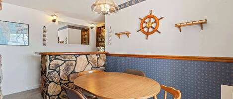 Un comedor con una mesa redonda de madera y sillas, una pared decorativa de piedra, un adorno con timón de barco y un espejo sobre una estantería.