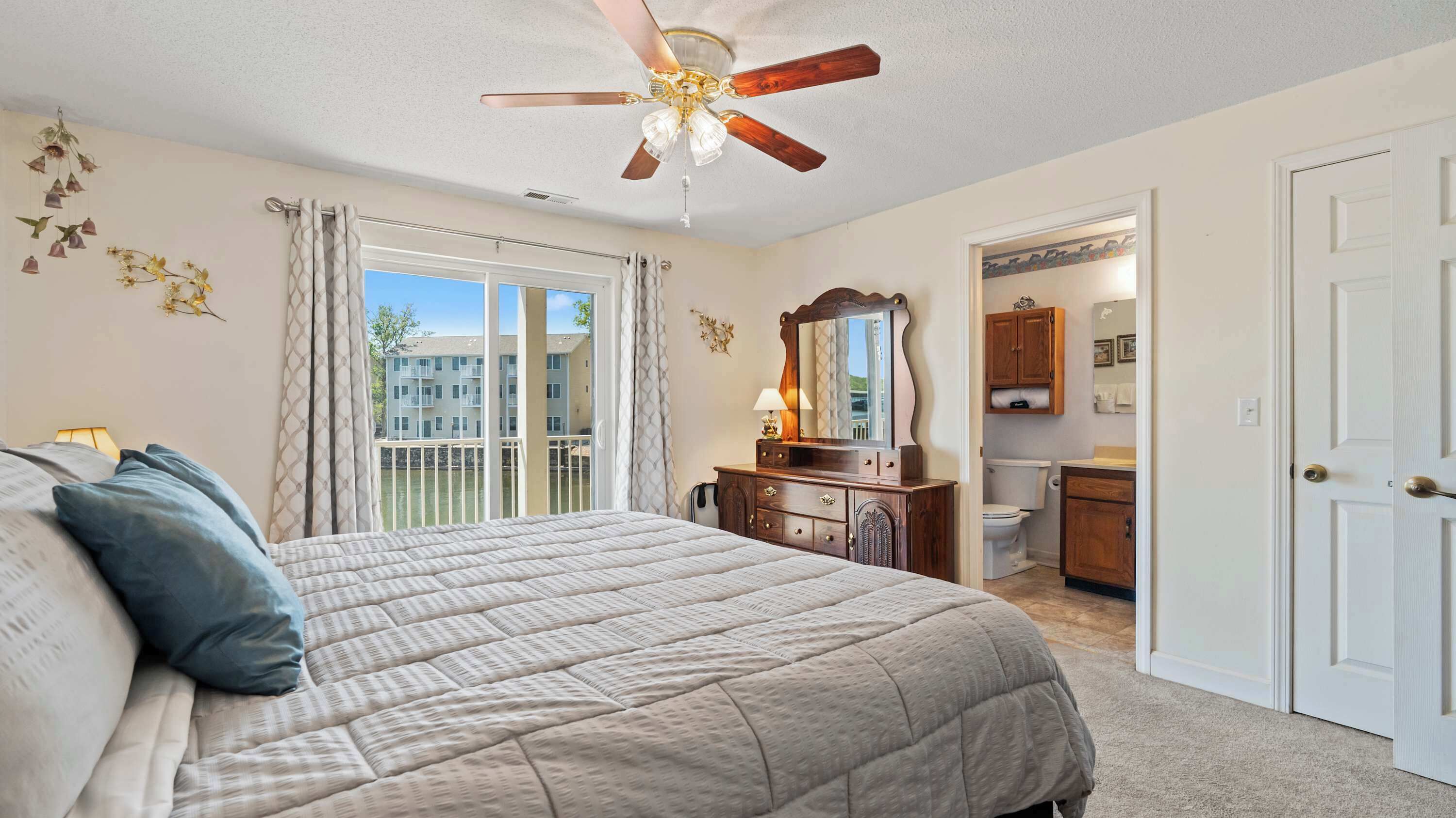 A bedroom with a bed, a ceiling fan, a dresser, and a view of a building through a window.