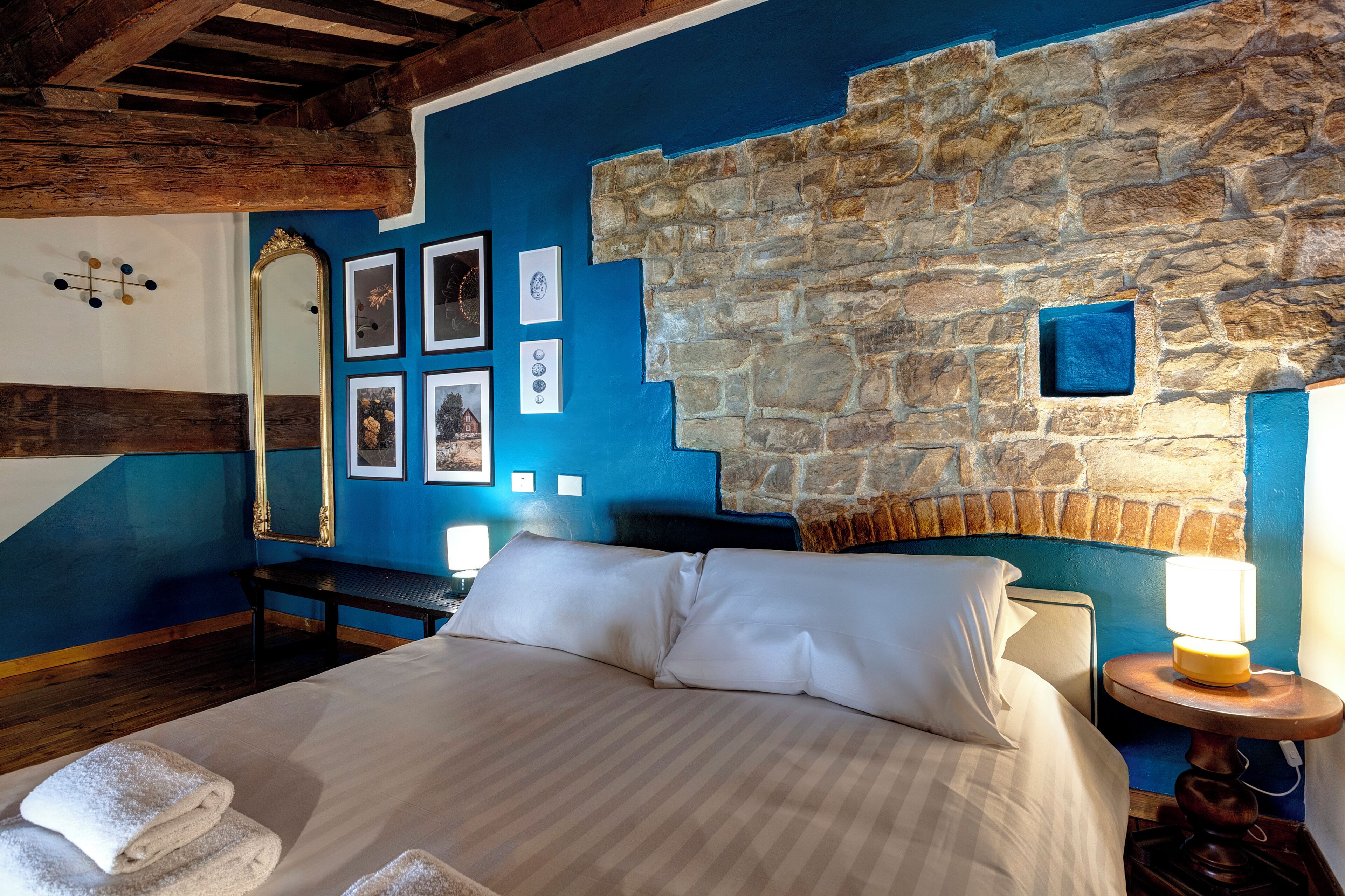 Un dormitorio con pared de piedra, cama con ropa blanca, mesita de noche con lámpara y cuadros enmarcados en la pared.