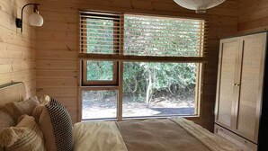 A wooden cabin room with a bed, a window with blinds, a wardrobe, and a hanging light fixture.