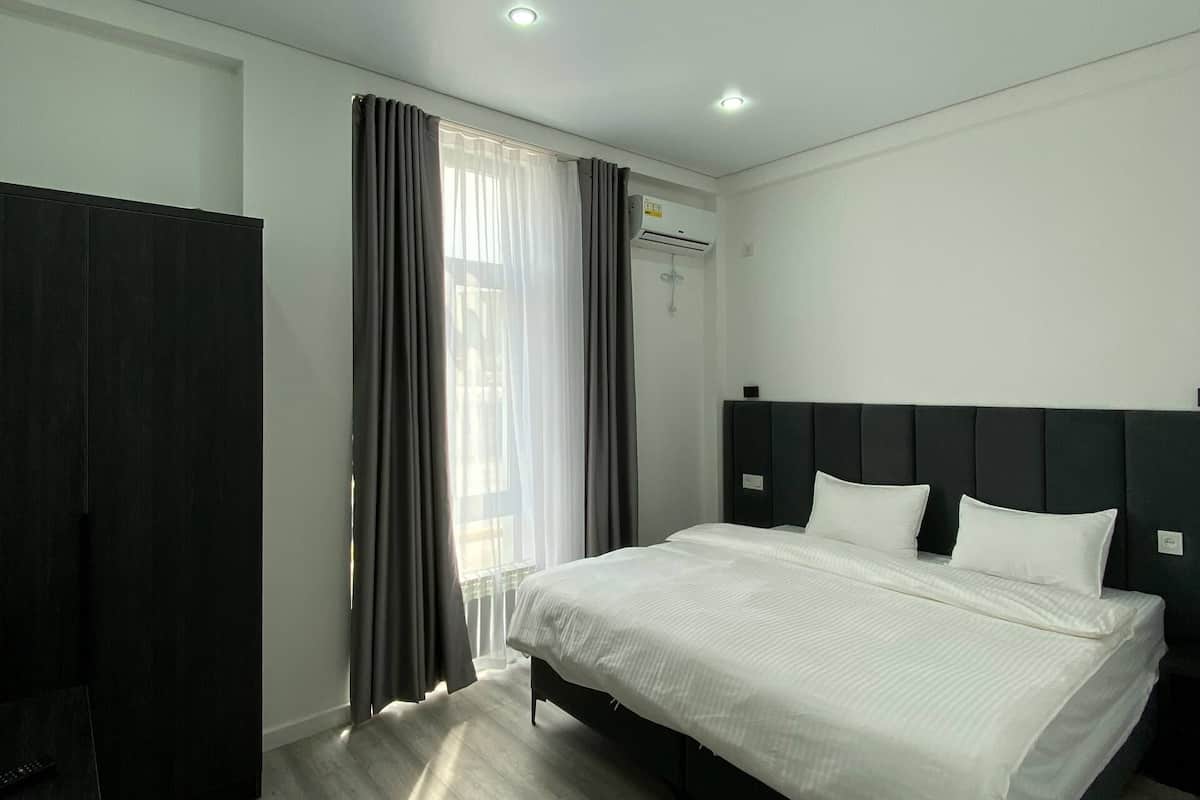 Habitación de hotel moderna con una cama grande, cabecero oscuro, suelo de madera y una ventana con cortinas.