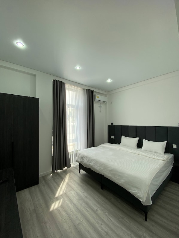 A modern hotel room with a large bed, dark headboard, wooden flooring, and a window with curtains.