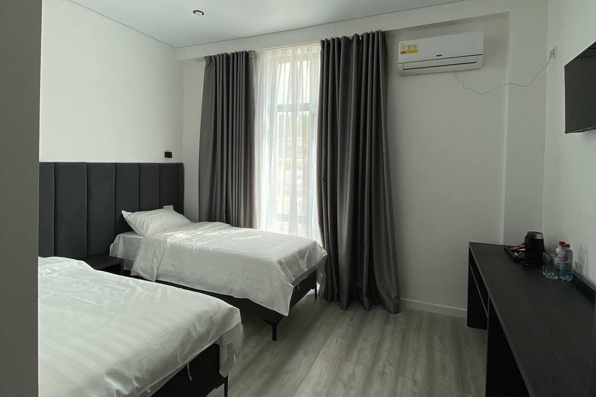 Habitación de hotel con dos camas, un escritorio, una ventana con cortinas, un equipo de aire acondicionado y un suelo de madera de color claro.