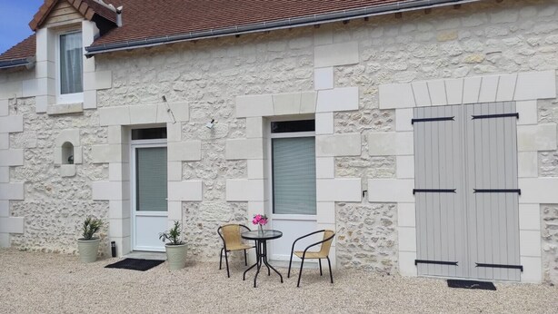 A stone building with a tiled roof, two windows with shutters, and a door. There is a small outdoor seating area with a table and chairs.