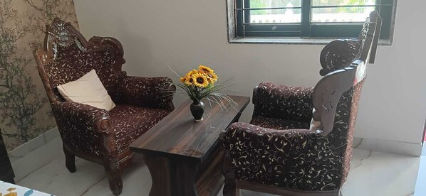 A room with a wooden chair and sofa set, a small table with a vase of flowers, and a window with a view. - Bohara Bhawan (Jaipur)