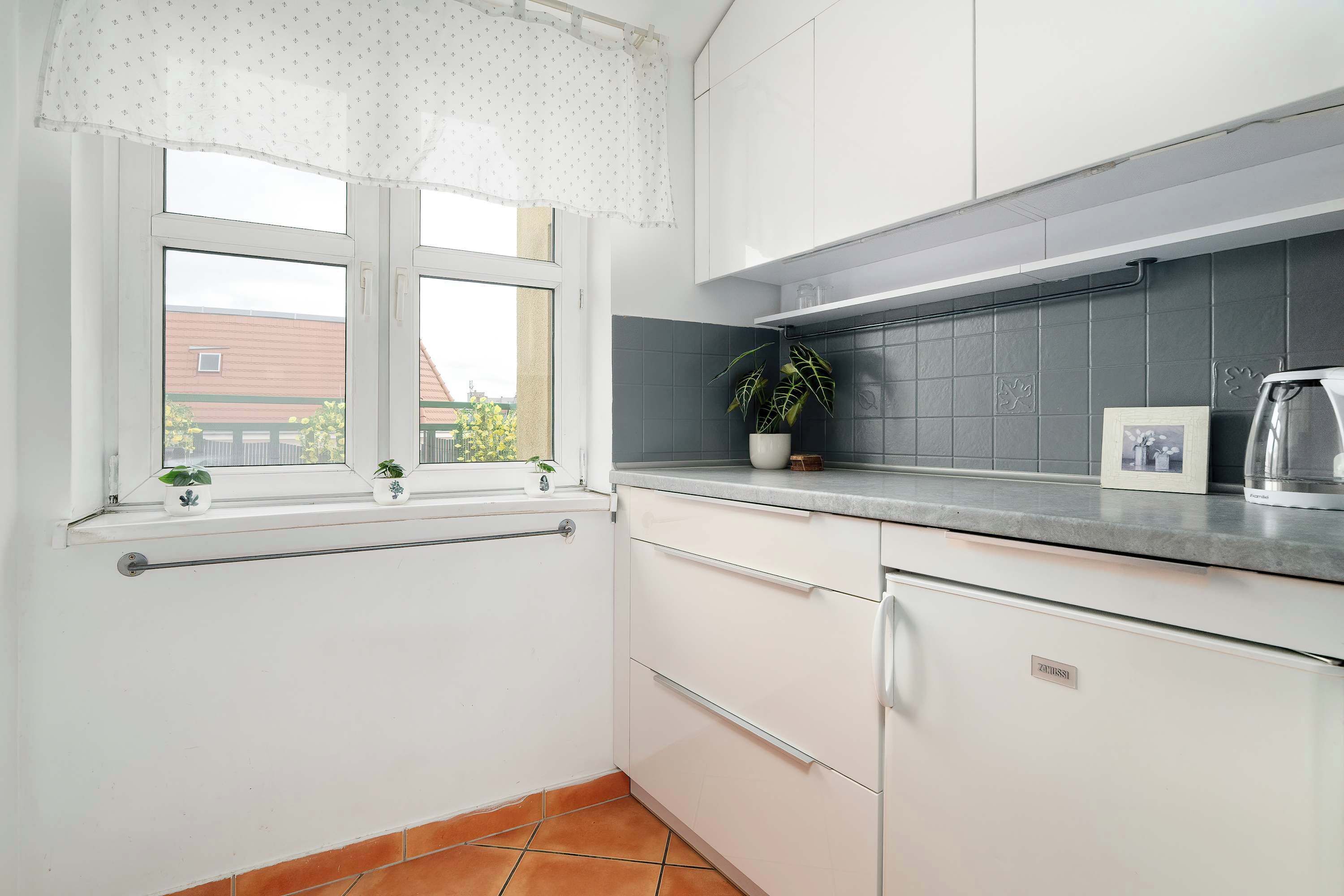 Eine Küche mit weißen Schränken, einem Fenster mit weißem Vorhang, einer Topfpflanze und einer gefliesten Spritzschutzwand.
