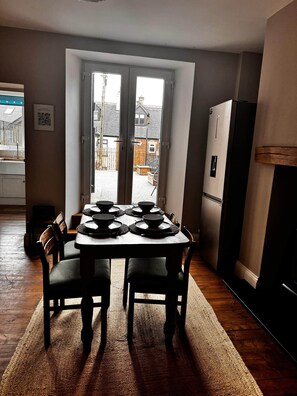 A dining area with a table set for four, a refrigerator, and a view of the outside through a door. - Cosy Buxton Terraced House (Buxton)