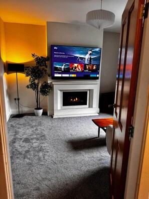 A living room with a flat-screen TV mounted above a fireplace, a potted plant, and a modern light fixture. - Cosy Buxton Terraced House (Buxton)