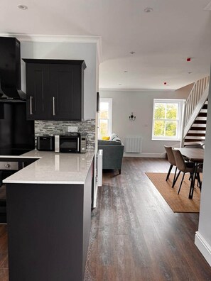 A modern kitchen with dark cabinets, a central island, and a dining area with a staircase. - Gorgeous Spacious Buxton Park Apartment (Buxton)