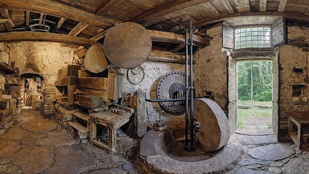 Un intérieur rustique avec des murs en pierre, une grande meule en pierre et une fenêtre donnant sur un paysage verdoyant.
