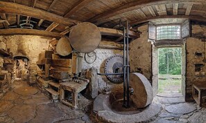 Un intérieur rustique avec des murs en pierre, une grande meule en pierre et une fenêtre donnant sur un paysage verdoyant.
