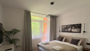 Un dormitorio con cama, ventana con cortinas, una planta y un cuadro en la pared.