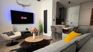 Una sala de estar moderna con un sofá gris, una mesa de centro, un televisor montado en la pared y un comedor visible a través de una puerta abierta.