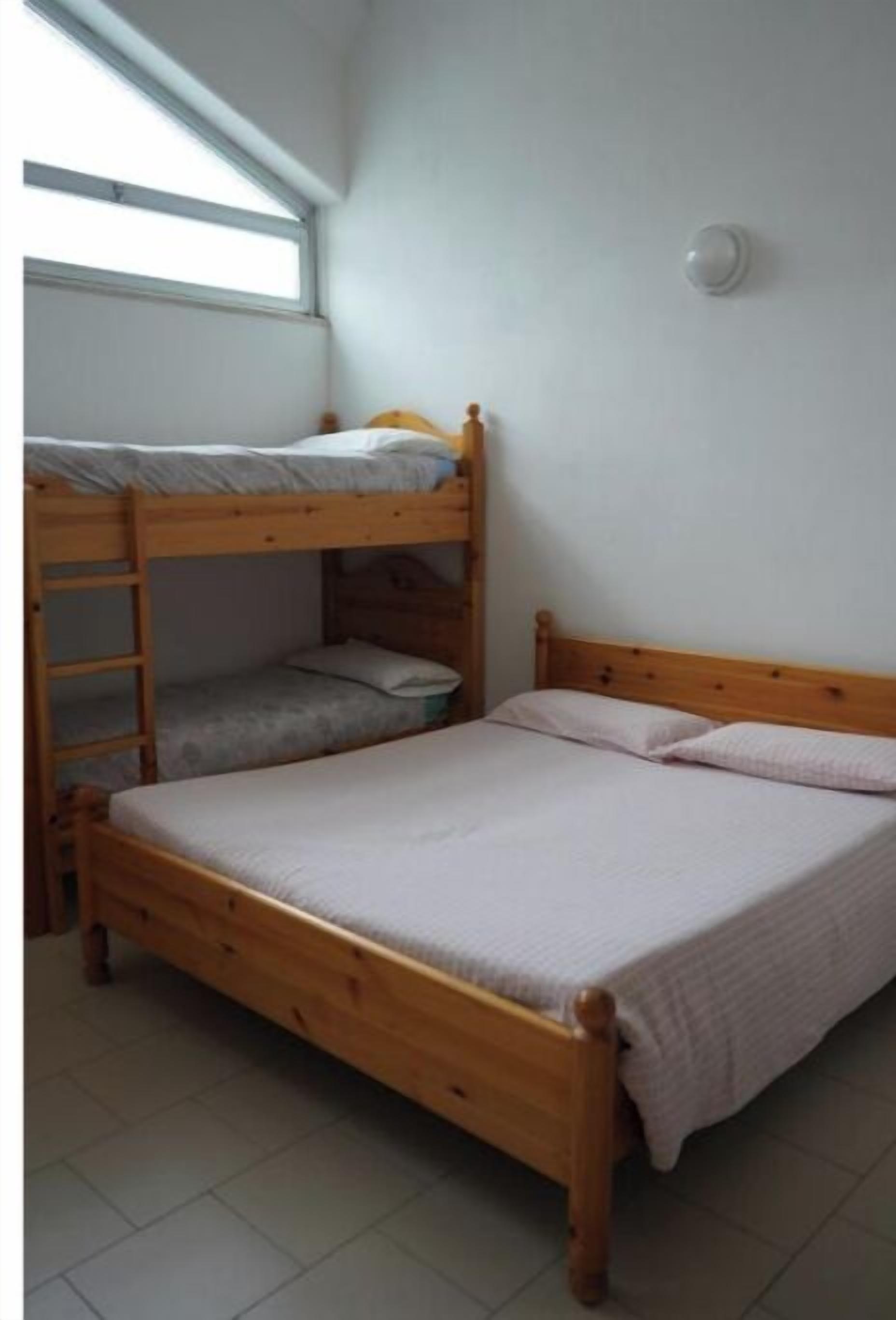 Une chambre avec un lit superposé, une fenêtre, une lampe fixée au mur et un sol carrelé.