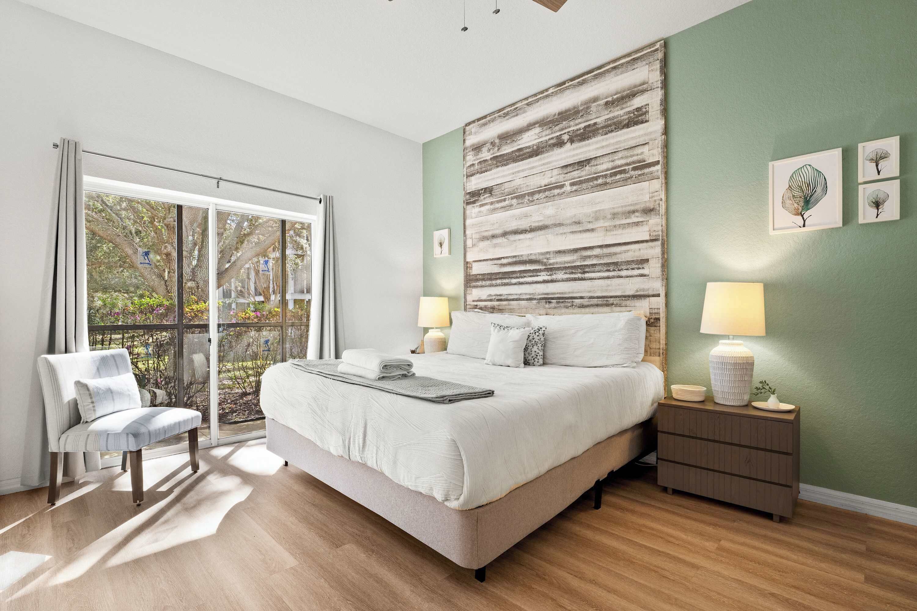 A bedroom with a large bed, a chair, a nightstand, and a view of the outdoors.