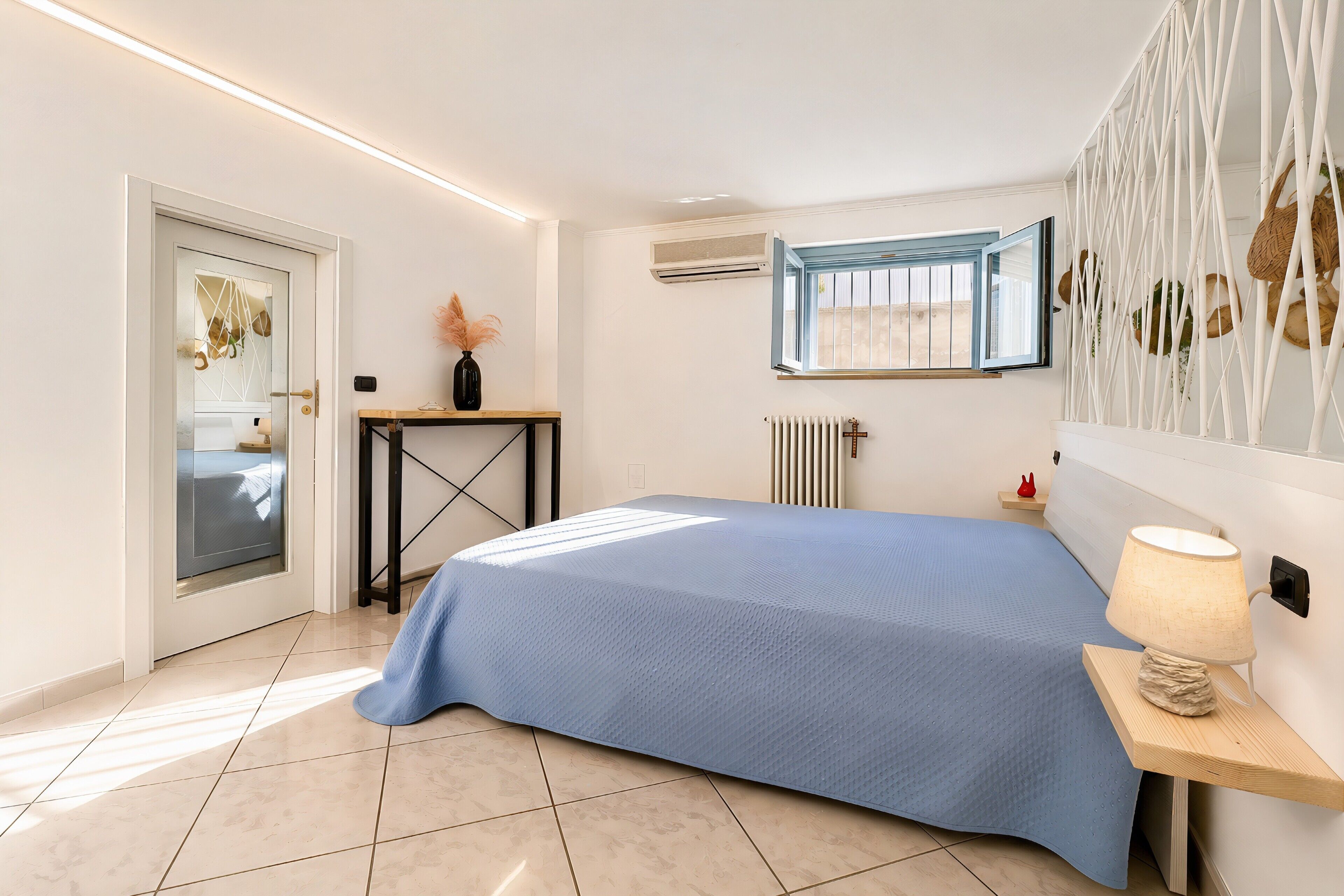 A bedroom with a bed, bedside table, lamp, window, radiator, and a door with a glass panel.